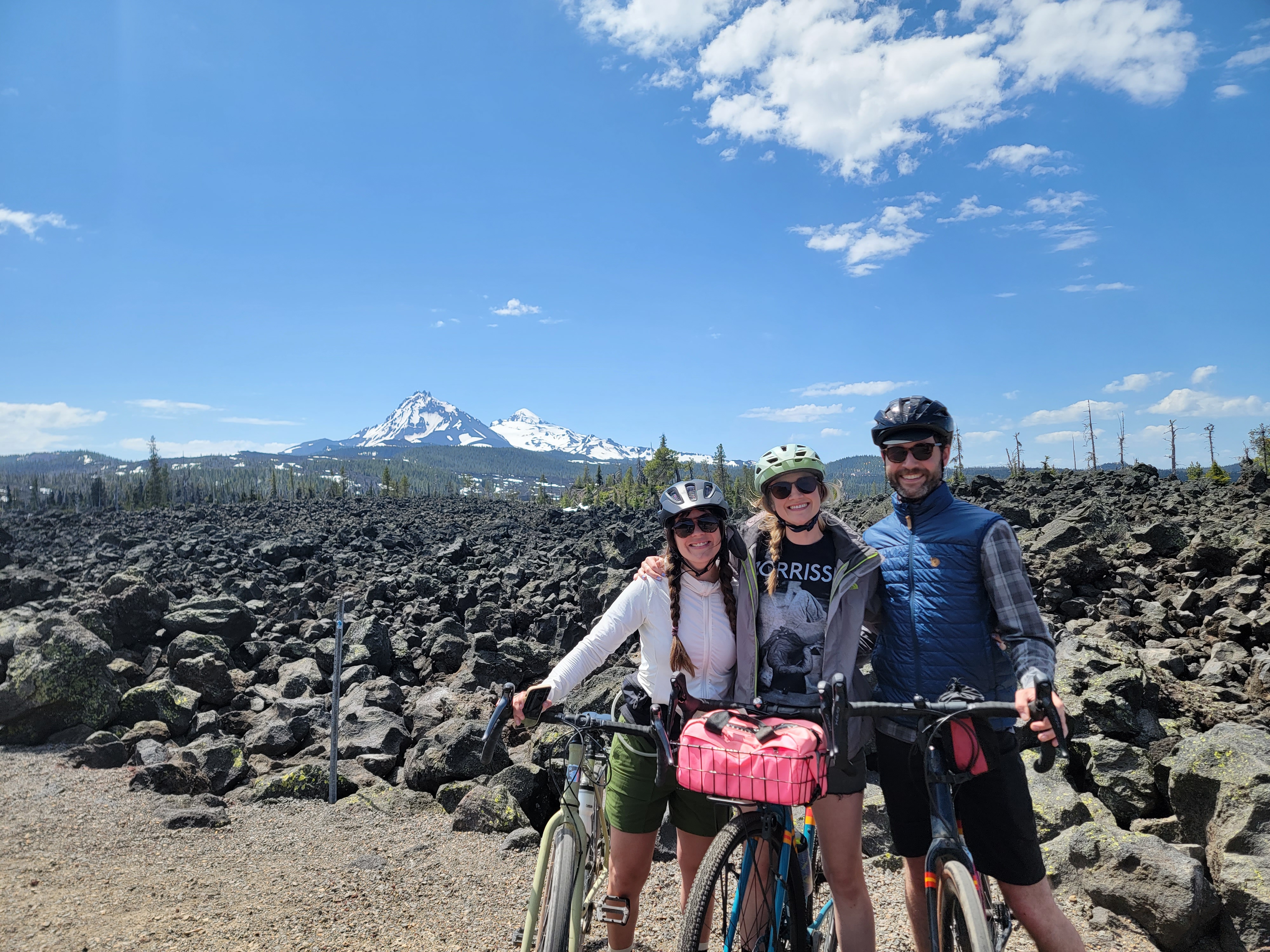 3 people with bikes posing at the summit of McKenzie Pass, sunny sky and mountain views in the background